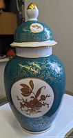 Full view of vase with lid showing teal body, gold scrollwork, floral cartouche
