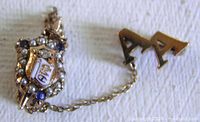 front view of Phi Delta Theta badge pin with seed pearls, sapphires, enamel shield and attached "Α Α" guard pin on chain