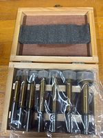 Open wooden box showing seven gold-tone Forstner bits sealed in plastic