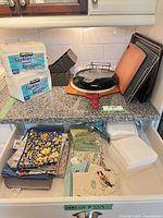 Overall lot on countertop and drawer showing napkin packs, bakeware stack, cutting boards, cloth linens