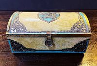 Front view of embossed metal chocolate box showing clasp and ornate scroll corners