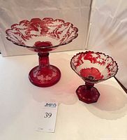 Two ruby-flashed cut-to-clear crystal pedestal bowls on white background; larger bowl left, smaller right; visible deer and forest designs