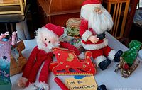 assorted Christmas decor on table: two Santa dolls, red felt sleigh, miniature metal sleigh, floral tin