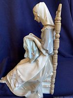 Left side view of entire figurine seated on chair holding cloth