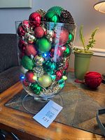 Full view of glass hurricane vase with ornaments, pinecones and lit string lights