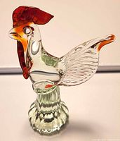 Front view of clear and green glass rooster with red-orange comb and tail on ribbed base