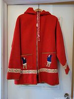 Full front view of red hooded wool coat showing zipper, pockets with appliqués and ribbon trim