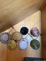 Seven hats displayed on wooden accordion rack against panel wall: straw sequin hat, tan felt hat, mustard velvet turban, pastel pink pillbox, black velvet pillbox, taupe velvet pillbox with red trim, olive green cloche