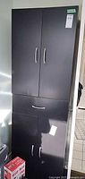 Front view of tall black cabinet showing upper doors, center drawer, lower doors, handles and overall condition