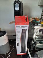 Lasko CT30796C boxed tower heater beside white cylindrical Dreo tower heater base