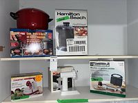Shelf with red pasta pot atop its NutriPro box, boxed Hamilton Beach juicer, Starfrit apple peeler box, white food grinder, Kenmore waffle maker box