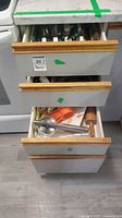 Three kitchen drawers open showing flatware, utensils and measuring tools