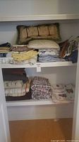 Closet shelves holding multiple folded sheets, blanket, decorative pillow and seat cushion