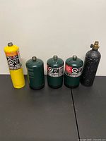All five empty gas cylinders lined up: yellow ProTorch MAP-Pro, three green Coleman propane, black unbranded tank