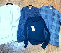 All four garments laid flat: sand cardigan, charcoal marled cardigan, black turtleneck, grey plaid cardigan