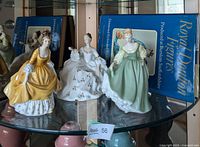 Three Royal Doulton porcelain lady figurines and Royal Doulton Figures book displayed on glass shelf