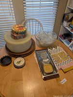 Overall lot view showing cake carrier on wood tray, tins stack, punch bowl, ashtrays, metal tray with contents, placemats