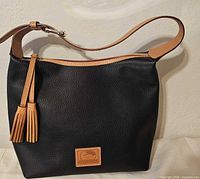 Front view of black pebble leather handbag showing tan trim, tassels, logo patch