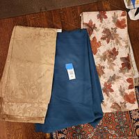 Shows three large rectangular tablecloths (gold damask, teal solid, cream with leaf print) on hangers
