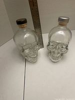 Front view of two clear glass skull bottles with ruler for scale