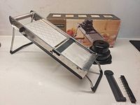 Mandoline on stand with hand guard, julienne comb and box visible