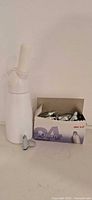 Whipped cream dispenser standing beside open charger box with loose cartridges visible