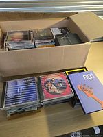 Open cardboard box filled with numerous CDs plus additional stacks on table