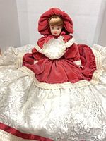 Full view of porcelain doll seated in red velvet dress with lace skirt