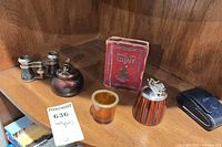 Group of six vintage items on shelf: opera glasses, two table lighters, red booklet, amber glass cup, leather pouch
