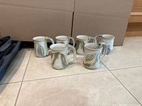 Six handmade stoneware mugs arranged on tiled floor showing glaze pattern and handles