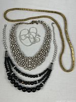 All four necklaces laid flat for full view: gold herringbone, silver snake chain, faux pearl rhinestone bib, black three-strand beads