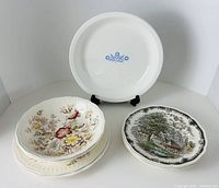 Front view showing all seven pieces: CorningWare pie plate on stand, stack of Montrose floral plates and bowls, rural-scene plate