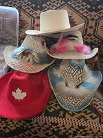Five hats stacked: red Canadian hat, three white straw Mexican hats with feather decoration, and one white felt Mexican hat on top