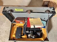 Open grey metal case showing camera, light meter, manuals and accessories in yellow foam interior