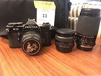 Front view of Pentax KX camera with attached 50 mm lens, two additional lenses, and bag
