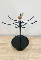 Front angle of black metal jewelry holder with gold bird finial, hooks and dish base visible