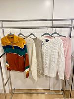 Five sweaters hanging on rack: multicolour quarter-zip, cream cardigan, white cable-knit, pink cable-knit