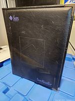 Front view of black leather portfolio showing Sun logo, embossed square, brass corner