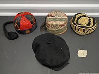 Four hats arranged on flat surface: red/black braided cap, woven fez, embroidered fez, black newsboy