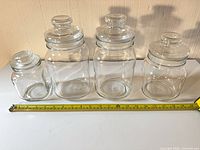 Four glass storage jars with lids lined up next to measuring tape