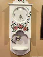 Front view of white floral wall clock showing dial, pendulum, painted roses