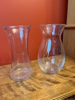 Two clear glass vases on wooden surface, one flared rim cylindrical shape, one bulbous