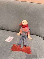 Duck figure seated on sofa showing front, wooden head, cloth clothing