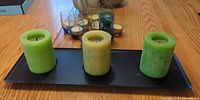Three pillar candles on black tray with coloured glass votive holders and two beige ceramics in background