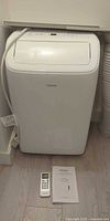 Front view of NOMA portable air conditioner with remote and manual on floor