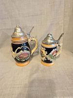 Front view of both ceramic beer steins showing scenic panels and pewter lids