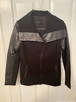 Front view of black Danier mixed-material jacket on hanger showing leather sleeves, textured leather shoulder panel, asymmetrical zipper, and pockets