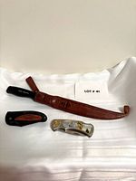 Overall view of three knives including Normark fillet knife in sheath, decorative folding knife, black/wood inlay folding knife