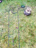 Group shot showing two tall black metal garden stakes, seven wire plant baskets, stack of round trays, and wood spool