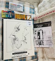 Assorted art supplies on plaid surface including sketchbook, oil pastels, coloured pencil set, drawing pad and rice paper roll